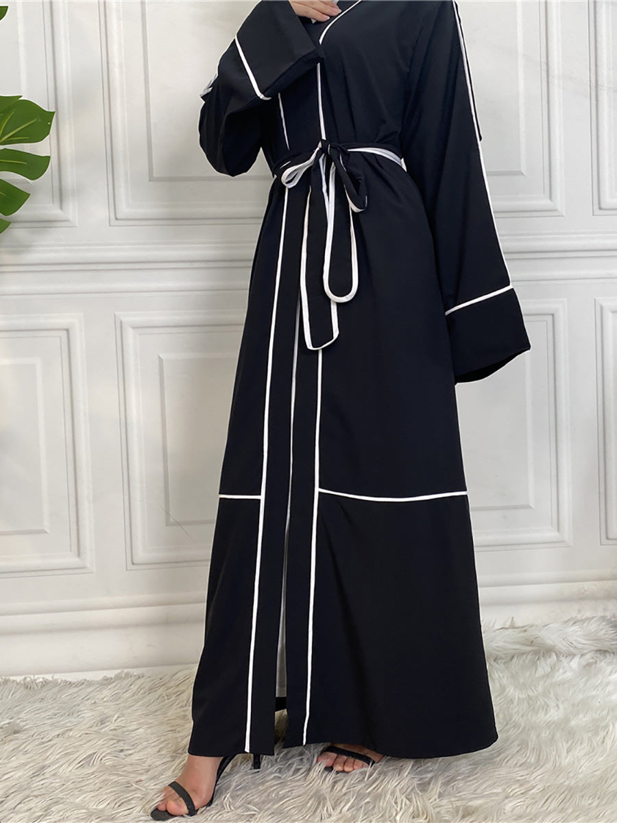 Person wearing a black abaya with white trim against a white paneled wall.